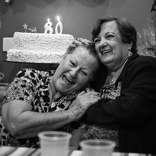 Two older women laughing and hugging with a birthday cake in the background with two burning candles in the shape of 8 and 0.