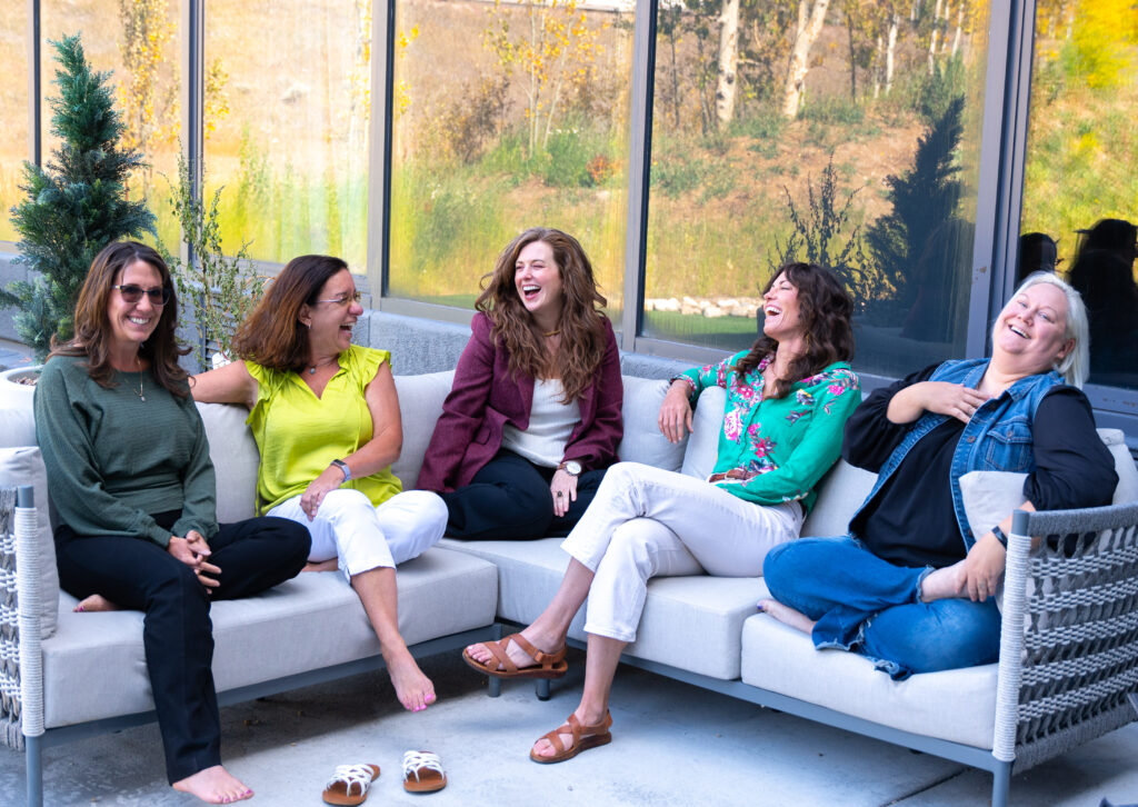 Vintage team including five women sitting on a grey couch laughing together.